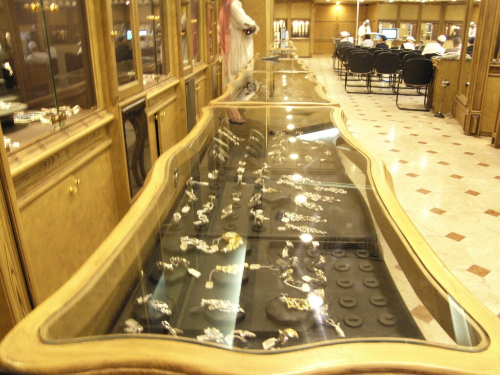 Interior photograph showcasing an elongated, curved wooden display counter filled with gold jewelry inside Makkah's largest showroom, with a dedicated auction seating area visible in the background